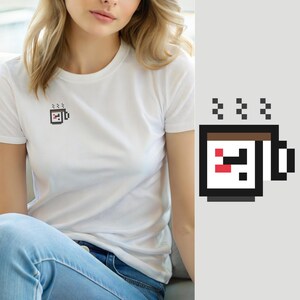 Op de afbeelding: Een wit T-shirt met korte mouwen en een kleine pixel art afbeelding op de linkerborst. De afbeelding toont een vierkante koffiemok met een zwarte omlijning, een bruine bovenkant en een klein rood accent, met drie pixelachtige stoomstreepjes.