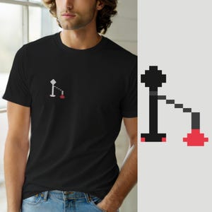 Puede incluir: Una camiseta negra de manga corta con un pequeño gráfico de pixel art en el pecho izquierdo. El diseño muestra un poste vertical negro con una parte superior circular, conectado por una línea diagonal a una base circular roja.