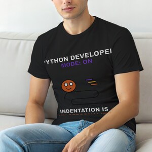 Puede incluir: Camiseta negra con el texto "PYTHON DEVELOPER MODE: ON" en blanco y morado. Debajo, una cara de dibujos animados y las palabras "INDENTATION IS" en blanco. La camiseta la lleva una persona.