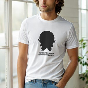 May include: White t-shirt with a black graphic of a brain and the text "ERROR 404 BRAIN UPDATE REQUIRED". The t-shirt has a crew neck and short sleeves. The person is wearing blue jeans.