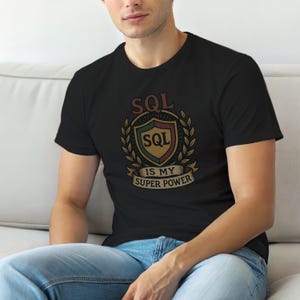 May include: A black short-sleeve t-shirt featuring a graphic design of a shield surrounded by laurel leaves. The text "SQL" appears above the shield and inside it, with a banner below the shield displaying the text "IS MY SUPER POWER".