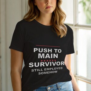 Puede incluir: Camiseta negra con texto blanco: "PUSH TO MAIN SURVIVOR STILL EMPLOYED SOMEHOW". El texto está apilado, con una línea roja y flechas encima de "SURVIVOR".