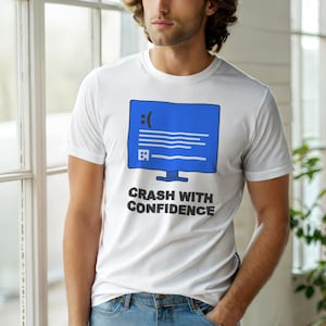 May include: White t-shirt featuring a blue computer screen graphic with a sad face and the text "CRASH WITH CONFIDENCE" in black. The design is centered on the front of the shirt.