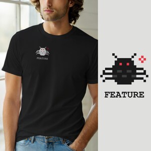Op de afbeelding: Een zwart T-shirt met korte mouwen en een kleine pixel-art afbeelding op de linkerborst. De afbeelding toont een donkergrijs insectachtig wezen met rode ogen en een rood kruissymbool, vergezeld door het woord FEATURE in witte hoofdletters.