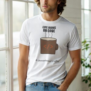 Op de afbeelding: Wit t-shirt met de tekst "LIFE RUNS ON CODE & Occasional Coffee". Een bruine koffiemok-afbeelding met codesymbolen staat op de voorkant. Het shirt is gemaakt van een zacht materiaal.