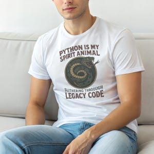 May include: A white short-sleeved t-shirt featuring a graphic of a coiled snake inside a circular border. The text reads "PYTHON IS MY SPIRIT ANIMAL" above the snake and "SLITHERING THROOUGH LEGACY CODE" below the snake in brown lettering.