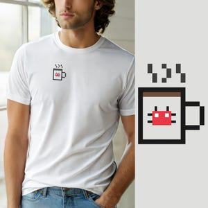 Op de afbeelding: Een wit T-shirt met korte mouwen en een kleine pixel art afbeelding op de linkerborst. De afbeelding toont een lichtgrijze mok met een bruine rand, een zwart handvat en een rood gepixeld krabicoon in het midden, met zwarte stoom erboven.