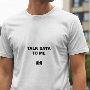 May include: White t-shirt with the text "TALK DATA TO ME" in black, along with a small graphic. The shirt is short-sleeved and appears to be made of a soft material. The person wearing the shirt is standing outside.