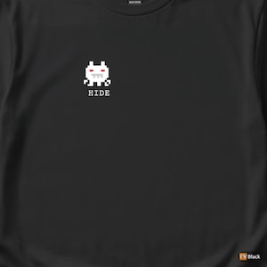 Puede incluir: Camiseta negra con un diseño de criatura blanca pixelada y detalles en rojo, sobre la palabra "HIDE" en blanco. El diseño está centrado. Hecha de algodón pesado.
