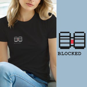 Peut inclure: Un t-shirt noir à manches courtes avec un petit graphique en pixel art sur la poitrine gauche. Le motif représente deux formes rectangulaires noires reliées par un petit carré rouge, avec le mot BLOCKED écrit en lettres capitales noires.