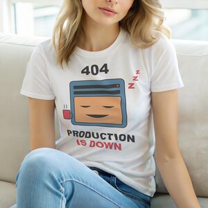 Może przedstawiać: Biała koszulka z grafiką z kreskówkowym ekranem komputera ze śpiącą twarzą, filiżanką kawy i napisem "404 PRODUCTION IS DOWN". Koszulka ma również napis "zzz" obok ekranu.