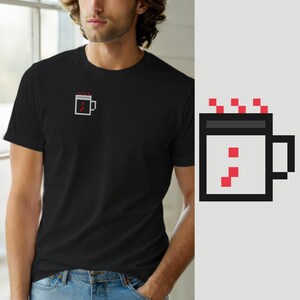 Può includere: Una t-shirt nera a maniche corte con una piccola grafica in pixel art sul petto sinistro. La grafica raffigura una tazza bianca con un bordo grigio scuro, un manico nero e vapore rosso pixelato che sale dalla parte superiore, con dettagli rossi.