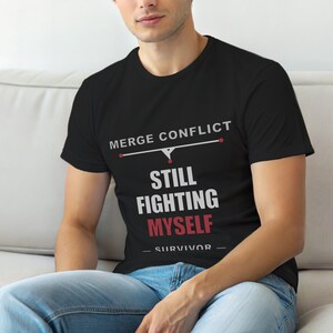 Puede incluir: Camiseta negra con texto blanco y rojo. El texto dice "MERGE CONFLICT" encima de "STILL FIGHTING MYSELF - SURVIVOR". La camiseta la lleva una persona sentada en un sofá blanco.