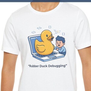 Op de afbeelding: Wit t-shirt met een afbeelding van een gele badeend op een laptopscherm, met een persoon en de tekst "Rubber Duck Debugging". Het ontwerp bevat codesymbolen.