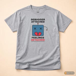 Könnte beinhalten: Graues T-Shirt mit dem Text "DEBUGGER ATTACHED" über einem blauen Robotergesicht mit X-Augen und einem gebrochenen Herzen. Darunter steht der Text "FEELINGS DETACHED" in Rot.