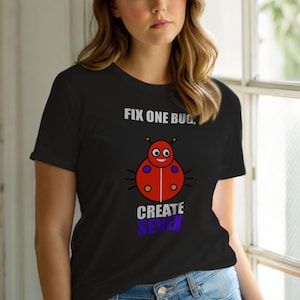 Op de afbeelding: Zwart T-shirt met een cartoon lieveheersbeestje ontwerp en de tekst "FIX ONE BUG, CREATE SEVEN." Het lieveheersbeestje is rood met blauwe en gele details. De tekst is in wit en paars.