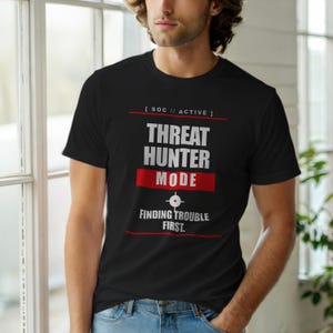 Op de afbeelding: Zwart T-shirt met witte en rode tekst met de tekst "THREAT HUNTER MODE" met de slogan "FINDING TROUBLE FIRST". Het shirt heeft ook de tekst "[ SOC // ACTIVE ]" boven de hoofdtekst. Het shirt wordt door een persoon gedragen.