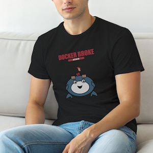 以下が含まれることがあります： 「DOCKER BROKE AGAIN」の赤い文字が入った黒いTシャツ。テキストの下には、メガネをかけた青いキャラクターの漫画の画像と笑顔があります。Tシャツは人が着ています。