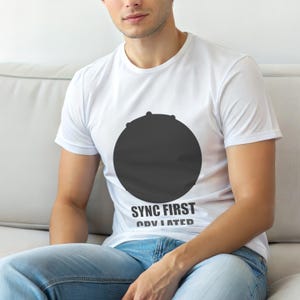 Puede incluir: Camiseta blanca con un gran diseño de círculo negro y el texto "SYNC FIRST OVULATED" en negro. La camiseta está hecha de un material suave.