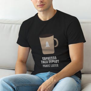 以下が含まれることがあります： ロケットが入ったコーヒーカップのグラフィックが特徴の黒いTシャツ。「ESPRESSO THEN DEPLOY PANIC LATER」というテキストがカップの下にあります。このシャツは柔らかい素材で作られています。