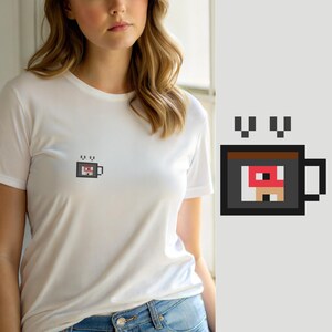 Pode incluir: Uma t-shirt branca de manga curta com um pequeno gráfico de arte de píxeis no peito. O gráfico mostra uma caneca de café cinzento-escuro com um rebordo castanho, contendo um design quadrado vermelho e bege, com duas pequenas nuvens de vapor.