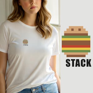 May include: A white short-sleeved t-shirt featuring a small, centered pixel-art graphic of a multi-layered burger with the word STACK printed in black capital letters directly underneath the illustration on the chest area.