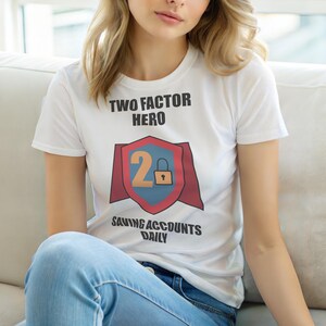 Op de afbeelding: Wit T-shirt met de tekst "TWO FACTOR HERO" boven een schildafbeelding. Het schild heeft een "2" en een slotpictogram, met de woorden "SAVING ACCOUNTS DAILY" eronder. De persoon draagt een blauwe spijkerbroek.