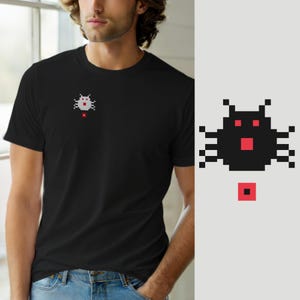 Może przedstawiać: Czarny t-shirt z krótkim rękawem z małą grafiką pixel art na lewej piersi. Grafika przedstawia stylizowane czarne stworzenie przypominające owada z czerwonymi oczami i czerwonymi kwadratowymi ustami, umieszczone nad pojedynczym czerwonym pikselem.