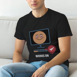 Könnte beinhalten: Schwarzes T-Shirt mit einem Computermonitor-Motiv, das ein lächelndes Gesicht zeigt. Das Design umfasst einen Fortschrittsbalken, ein Häkchen-Abzeichen und den Text "WORKS ON" unter dem Monitor.