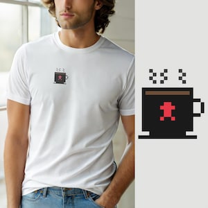 Könnte beinhalten: Ein weißes T-Shirt mit kurzen Ärmeln und einer kleinen Pixel-Art-Grafik auf der linken Brust. Die Grafik zeigt eine schwarze Kaffeetasse mit braunem Rand, aufsteigendem Dampf und einem kleinen, roten, verpixelten Lebkuchenmann auf der Vorderseite.