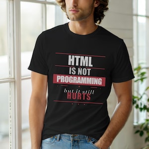 Op de afbeelding: Zwart T-shirt met de tekst "HTML IS NOT PROGRAMMING but it still HURTS" in wit en rood. Het shirt wordt gedragen door een persoon voor een raam.