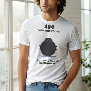 May include: White t-shirt with the text "404 PAGE NOT FOUND" above a black, round graphic with eyes and small lines. Below the graphic, the text reads "BUT BUGS FOUND EVERYWHERE". The shirt is worn by a person.