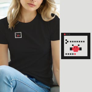 Peut inclure: Un t-shirt noir à manches courtes avec un petit motif carré sur la poitrine gauche. Le motif représente une fenêtre de terminal informatique gris clair avec une bordure noire, contenant une icône de bug rouge et du texte en pixels noirs.