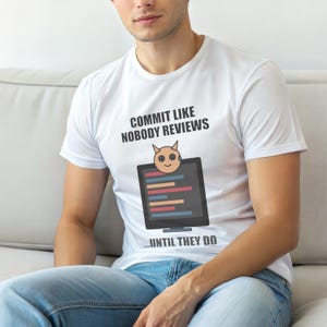 Op de afbeelding: Wit T-shirt met de tekst "COMMIT LIKE NOBODY REVIEWS" boven een cartoonafbeelding van een computerscherm met een lachend aliengezicht en de tekst "...UNTIL THEY DO". Het T-shirt wordt door een persoon gedragen.