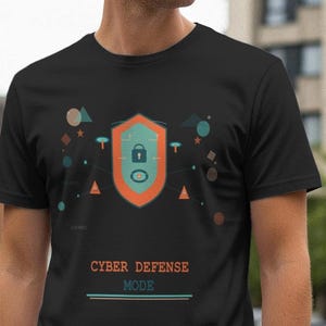 Può includere: T-shirt nero con un design a scudo, un'icona a forma di lucchetto e forme geometriche in verde acqua, arancione e marrone. Il testo "CYBER DEFENSE MODE" è stampato in arancione. La maglietta è a maniche corte.