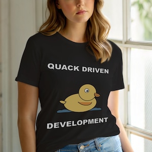 Op de afbeelding: Zwart T-shirt met de witte tekst "QUACK DRIVEN DEVELOPMENT". Een cartoon eend drijft op een blauwe waterlijn. De eend is geel met een oranje snavel en een enkel oog.