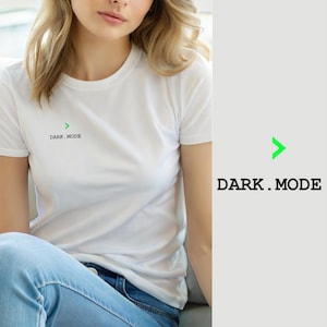 Könnte beinhalten: Ein weißes kurzärmeliges T-Shirt mit einem kleinen grünen Chevron-Symbol über dem schwarzen Schriftzug "DARK.MODE" auf der linken Brustseite. Das T-Shirt hat einen Rundhalsausschnitt und wird mit einer blauen Jeans kombiniert.