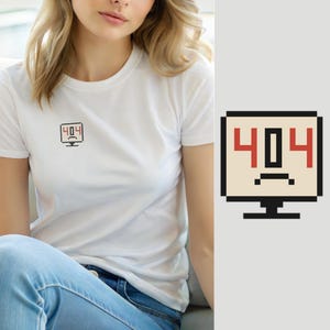 Op de afbeelding: Een wit T-shirt met korte mouwen en een kleine, gepixelde afbeelding op de borst. De afbeelding toont een computermonitor in zwart, beige en rood, met de tekst 404 en een droevig, fronsend gezichtssymbool onder de cijfers.
