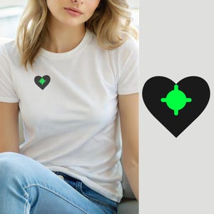 Może przedstawiać: Biały t-shirt z krótkim rękawem z małą grafiką na lewej piersi. Grafika składa się z czarnego serca z jaskrawozielonym symbolem celownika umieszczonym centralnie wewnątrz, co tworzy projekt o wysokim kontraście.