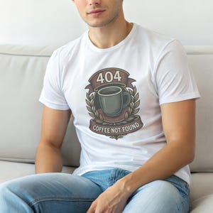Könnte beinhalten: Ein weißes Kurzarm-T-Shirt mit einer zentralen Grafik einer dunkelgrünen Kaffeetasse, die von einem Lorbeerkranz umgeben ist. Über der Tasse steht der Text 404 und unter der Tasse befindet sich ein Banner mit dem Text COFFEE NOT FOUND in Dunkelrot und Beige.