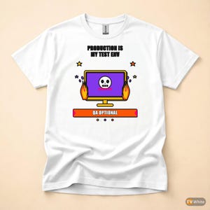 Op de afbeelding: Wit T-shirt met een grafisch ontwerp met een computermonitor met een schedel, vlammen en de tekst "PRODUCTION IS MY TEST ENV" en "QA OPTIONAL". Het ontwerp bevat sterren en een paars en oranje kleurenschema.