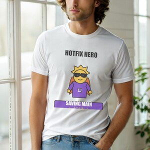 Puede incluir: Camiseta blanca con el texto "HOTFIX HERO" sobre un sol de dibujos animados con gafas de sol y capa. Debajo del sol hay una pancarta morada que dice "SAVING MAIN AGAIN".