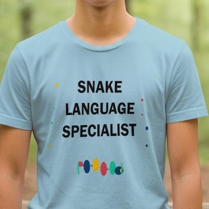 Op de afbeelding: Lichtblauw T-shirt met de zwarte tekst "SNAKE LANGUAGE SPECIALIST". Het shirt heeft een ronde hals en korte mouwen. Daaronder staan kleurrijke, abstracte vormen. De achtergrond is een vage bosscène.
