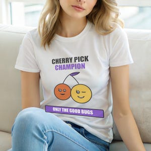 Puede incluir: Camiseta blanca con el texto "CHERRY PICK CHAMPION" en negro y morado. Debajo, un diseño de cerezas de dibujos animados con dos caras sonrientes. El texto "ONLY THE GOOD BUGS" está debajo.