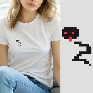Pode incluir: Uma t-shirt branca de manga curta com um pequeno gráfico pixelizado de uma cobra preta com olhos vermelhos e língua vermelha na zona do peito esquerdo. O design da cobra é composto por blocos quadrados, criando uma estética digital retro.