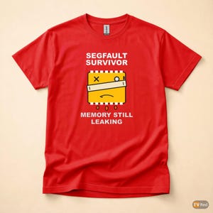 Op de afbeelding: Rood t-shirt met de tekst "SEGFAULT SURVIVOR" boven een gele afbeelding van een computercomponent met een verdrietig gezicht, een X en een witte balk. Daaronder staat de tekst "MEMORY STILL LEAKING".