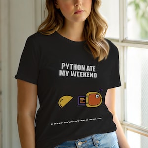 Puede incluir: Camiseta negra con el texto blanco "PYTHON ATE MY WEEKEND". El gráfico presenta un ordenador y un emoji naranja durmiendo. La parte inferior de la camiseta tiene más texto.