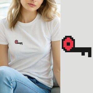 Może przedstawiać: Biały t-shirt z krótkim rękawem z małą grafiką pixel art na piersi. Grafika przedstawia czarny klucz z czerwoną, okrągłą główką i kwadratowym otworem pośrodku, wykonany w blokowym, cyfrowym stylu nawiązującym do gier wideo w stylu retro.