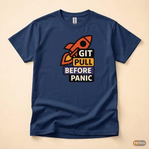 Op de afbeelding: Marineblauwe t-shirt met een raket-afbeelding en de tekst "GIT PULL BEFORE PANIC". De raket is oranje en zwart. Het t-shirt heeft een ronde hals en korte mouwen.