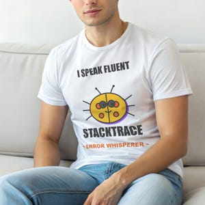 Op de afbeelding: Wit T-shirt met de tekst "I SPEAK FLUENT STACKTRACE" en een cartoon bug graphic. De bug is geel met zwarte antennes en een paarse omtrek. Onder de bug staat de tekst "ERROR WHISPERER".